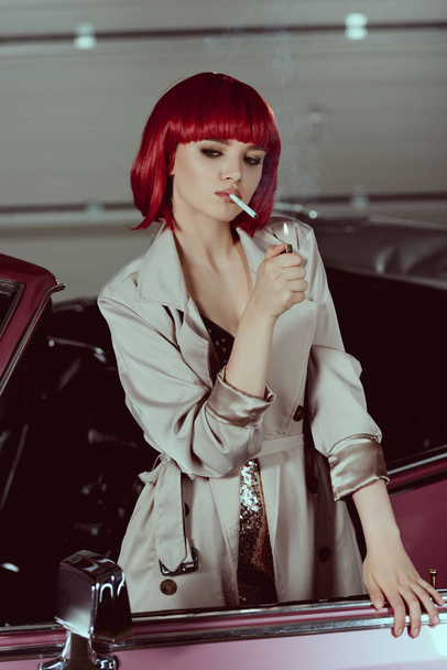 beautiful young woman in red wig and stylish trench coat smoking cigarette in classic car  - 写真・画像