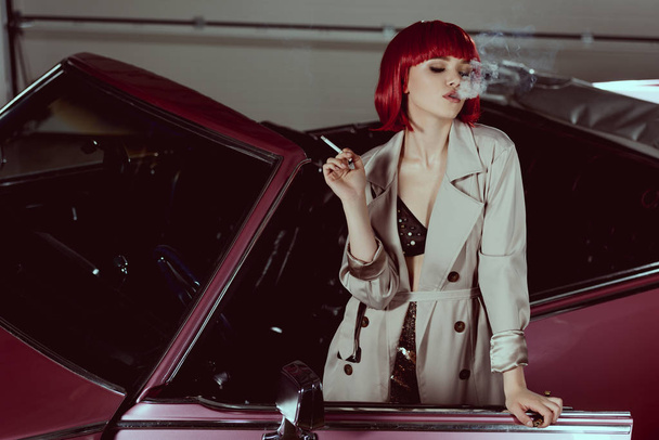 stilvolles Mädchen mit roter Perücke und Trenchcoat, das in einem Oldtimer Zigarette raucht  - Foto, Bild