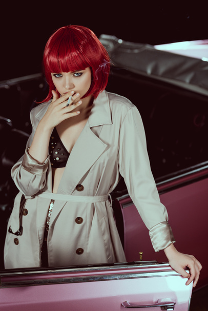 belle femme en perruque rouge et trench coat fumer cigarette et regarder la caméra près de la voiture
 - Photo, image