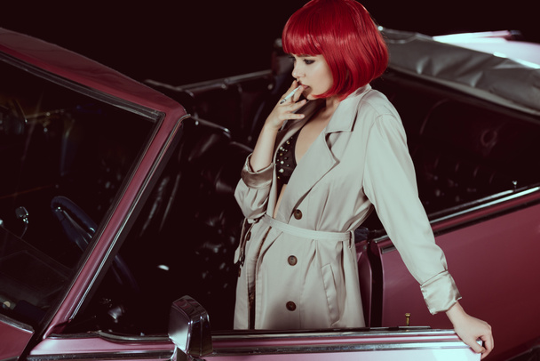 vista de ángulo alto de la chica con estilo en peluca roja y gabardina fumar cigarrillo cerca de coche vintage
 - Foto, Imagen