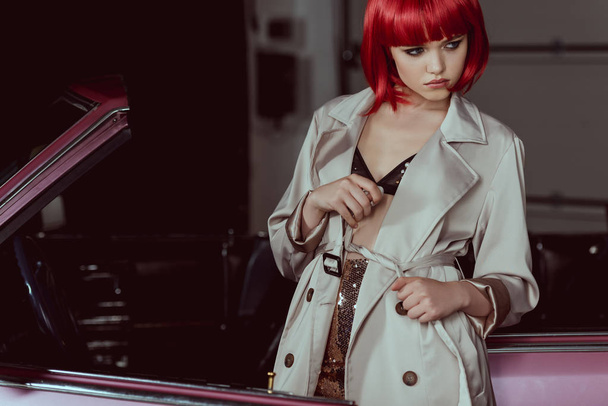 stylish girl in red wig tying trench coat and looking away near car - Foto, Imagen