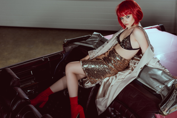 high angle view of stylish girl in red wig and trench coat looking at camera while sitting on retro car - Foto, Imagen