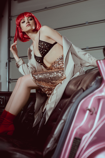 low angle view of stylish girl in red wig looking away while sitting on vintage car - 写真・画像