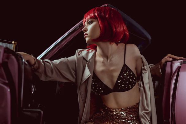 vista de ángulo bajo de la chica en peluca roja y gabardina mirando hacia otro lado mientras está sentado en el coche vintage
 - Foto, Imagen