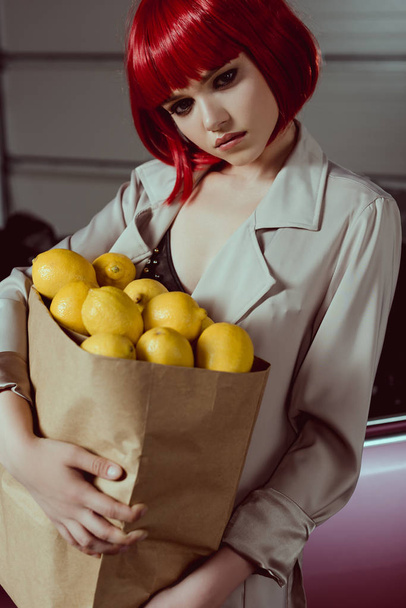 pensive girl in red wig and stylish trench coat holding paper bag with lemons - 写真・画像