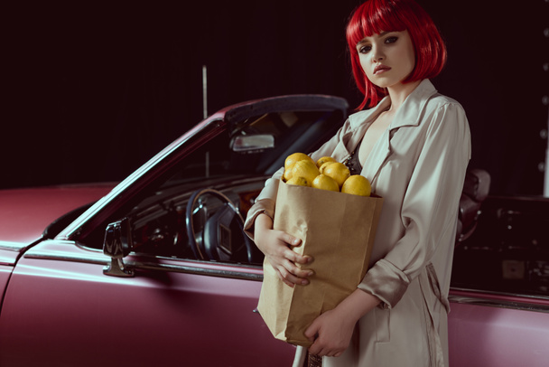 Frau in roter Perücke und schickem Trenchcoat hält Papiertüte mit Zitronen in der Hand und blickt in die Kamera, während sie in der Nähe des Retro-Autos steht   - Foto, Bild