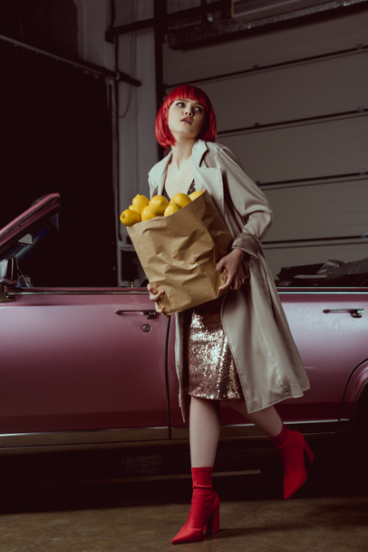 woman in red wig and stylish trench coat holding paper bag with lemons near retro car   - Φωτογραφία, εικόνα