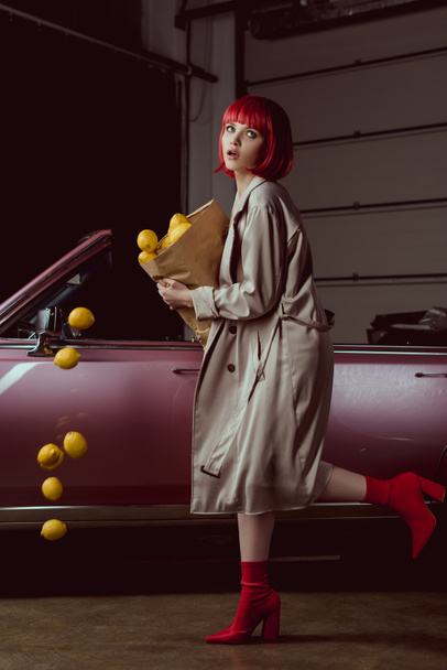 girl in red wig and stylish trench coat holding paper bag with falling lemons near retro car  - Foto, afbeelding