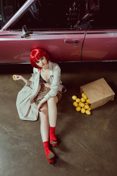 high angle view of beautiful girl in stylish trench coat holding lemon and looking at camera while sitting near retro car  - Foto, Imagem