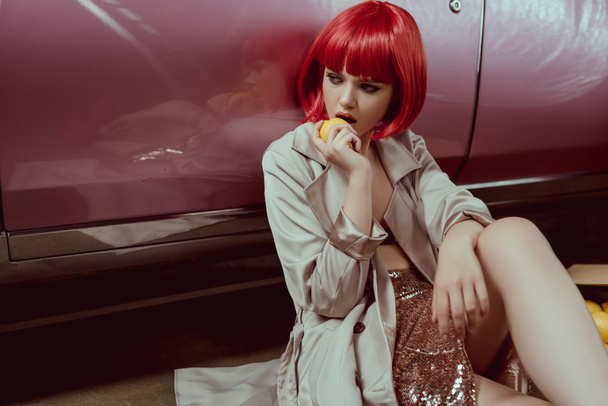 mujer joven con estilo en peluca roja comer limón mientras está sentado cerca de coche retro
 - Foto, Imagen
