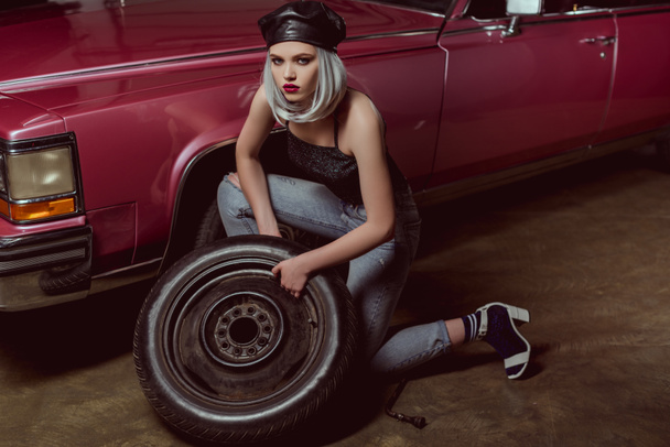 high angle view of beautiful stylish blonde girl in leather beret repairing car wheel and looking at camera - Foto, afbeelding