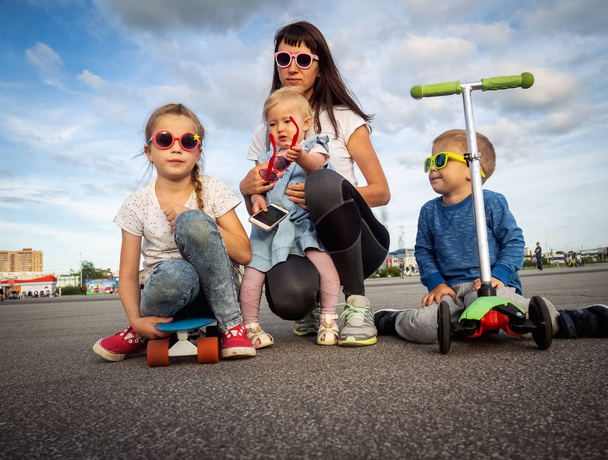 Foto móvil: Mamá y tres niños en gafas de sol con un monopatín y un scooter están sentados en el asfalto
. - Foto, Imagen