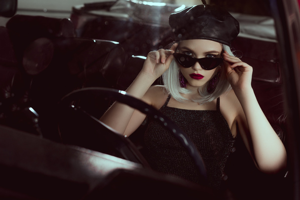 beautiful fashionable blonde girl in beret and sunglasses sitting in car and looking at camera - 写真・画像
