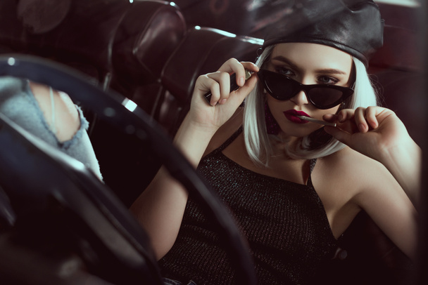 beautiful stylish blonde girl in beret and sunglasses holding cigarette while sitting in car - Photo, Image