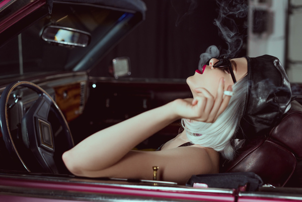 side view of sensual blonde woman in sunglasses and beret smoking cigarette in retro car  - Photo, Image
