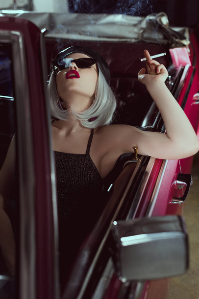 stylische blonde Frau mit Sonnenbrille und Baskenmütze, die Zigarette raucht, während sie im Retro-Auto sitzt  - Foto, Bild