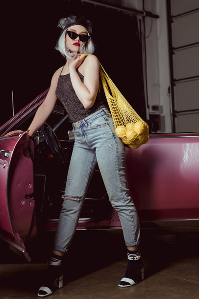 full length view of stylish blonde girl in beret and sunglasses holding string bag with lemons while standing near old-fashioned automobile - Foto, Imagen