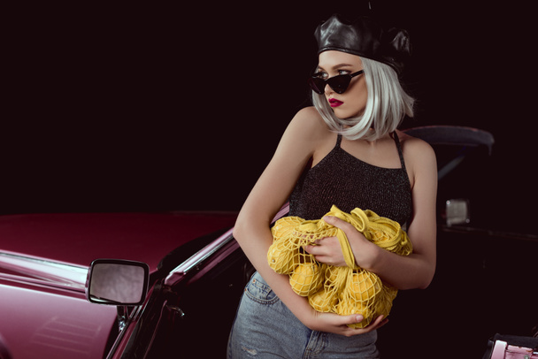 beautiful blonde woman in sunglasses and beret holding string bag with lemons and looking away while standing near car  - Foto, afbeelding