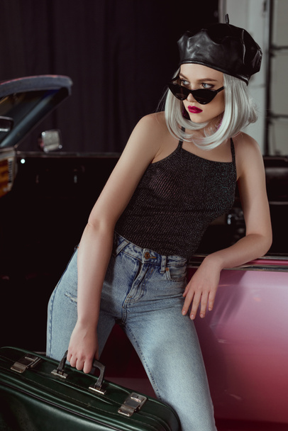 fashionable blonde girl in beret and sunglasses leaning at car and holding vintage suitcase - Foto, afbeelding