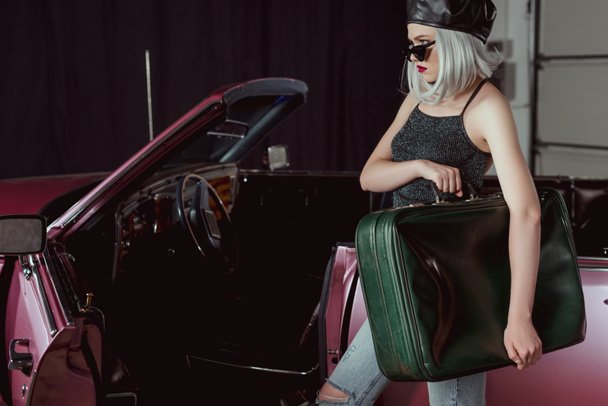Stílusos szőke lány strandpapucsot és gazdaság retro bőrönd közelében gesztenyebarna autó állva beret  - Fotó, kép