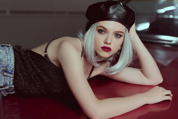 beautiful blonde woman in black beret lying on maroon car and looking at camera - Foto, imagen