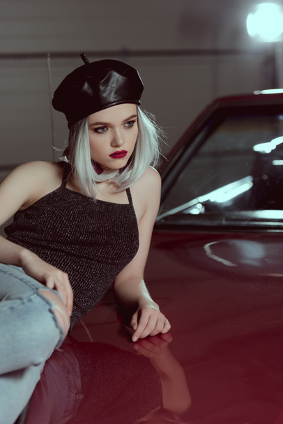 mooie blonde vrouw in zwarte baret liggend op kastanjebruine auto en de weg op zoek   - Foto, afbeelding