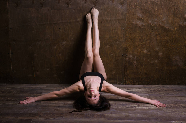 Theme is a sport and a beautiful sexy woman. Young caucasian girl lying on the floor kicking up posing in a black sports swimsuit clothes with bare feet on a dark wooden background in the studio. - Foto, imagen