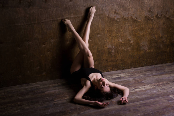 Theme is a sport and a beautiful sexy woman. Young caucasian girl lying on the floor kicking up posing in a black sports swimsuit clothes with bare feet on a dark wooden background in the studio. - Zdjęcie, obraz