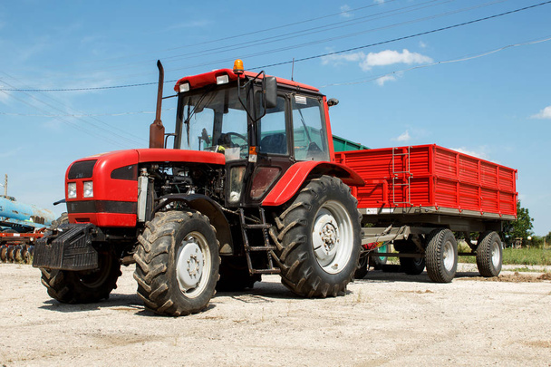 Czerwony Traktor z przyczepą zaparkowany na gospodarski przeciw błękitne niebo, słoneczny letni dzień - Zdjęcie, obraz