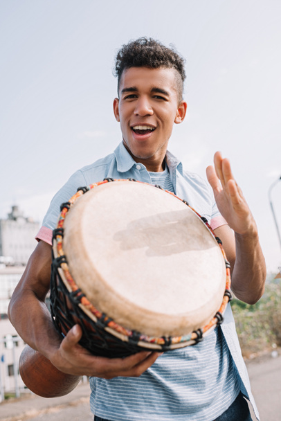 Jong en gelukkig Afrikaanse Amerikaanse straat muzikant met djembe uitvoeren op zonnige stad straat - Foto, afbeelding