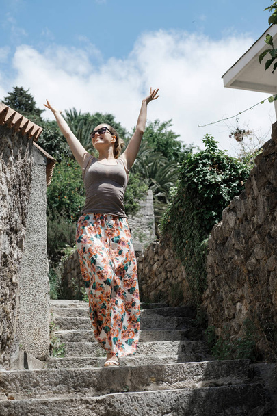 νεαρή γυναίκα τουριστικές βόλτες στην παλιά πόλη στο Μαυροβούνιο. Ευτυχισμένος κορίτσι σε γυαλιά ηλίου απολαμβάνει τη θερινή ώρα - Φωτογραφία, εικόνα