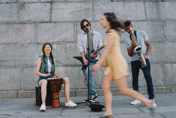 Mujer pasando por banda multirracial de músicos callejeros en entorno urbano
 - Foto, Imagen