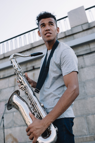 Jong en gelukkig Afrikaanse Amerikaanse straat muzikant bedrijf saxofoon in stad - Foto, afbeelding