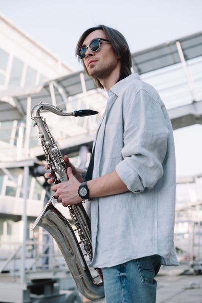Elegante chico con gafas de sol sosteniendo saxofón en la ciudad
 - Foto, Imagen