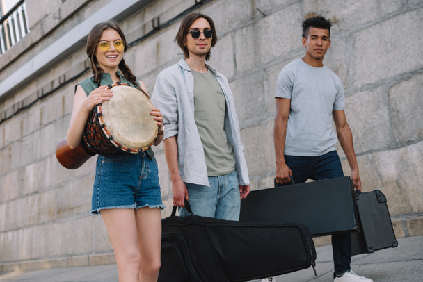 Chica joven y hombres multirraciales caminando y llevando instrumentos musicales en la soleada calle de la ciudad
 - Foto, Imagen