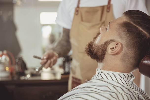 Man with a beard waits for a shave with a razor in a barbershop. Photo in vintage style - Foto, afbeelding