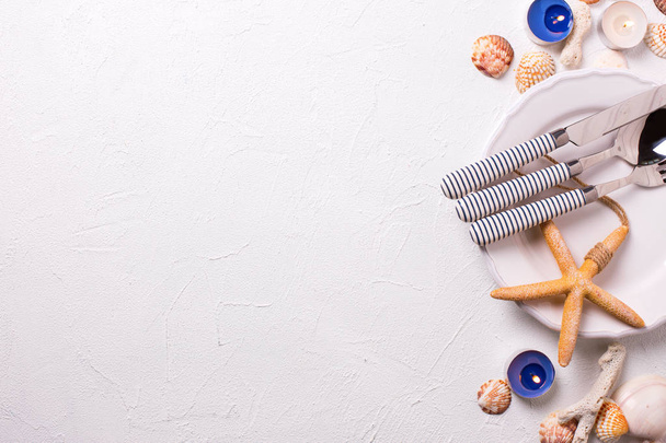 Summer table setting. Border from nautical  elements and white plate on white textured  background. Shells, cutlery, candles, sea star, coral. View from above. Selective focus. Place for text. - Φωτογραφία, εικόνα