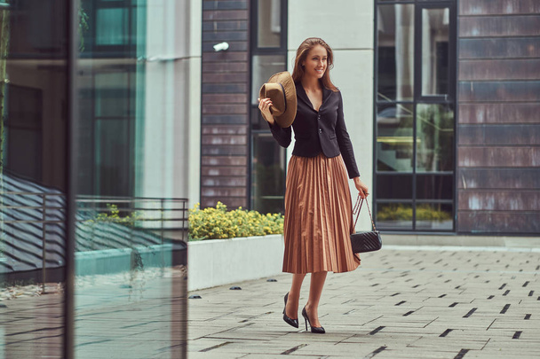 Mujer elegante moda feliz con una chaqueta negra, sombrero marrón y falda con un bolso de embrague caminando por el centro de la ciudad europea
. - Foto, imagen