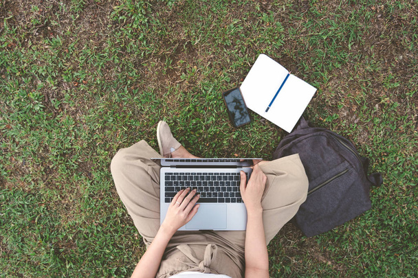 Top view of young freelancer woman using laptop in garden with smartphone, bag, notebook and pencil for working. Happy girl blogger writes blog content in outdoor park. Lifestyle working and device. - Foto, Imagem