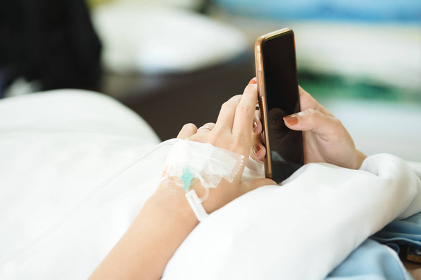 Close up woman patient hand with IV set using smartphone contact to doctor, emergency call service while lying on bed. Alone sick female chatting and social network with friend, family in hospital - Photo, Image