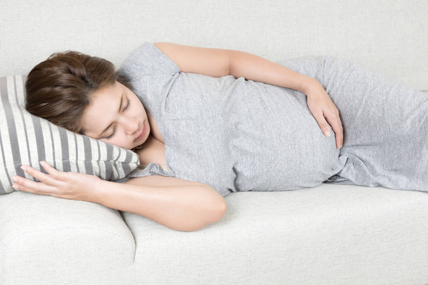 Young beautiful pregnant woman sleeping on gray sofa and pillow at home. Happy asian expectant laying on bed for rest and hand touching her belly. - Photo, Image