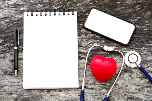 Health red heart with stethoscope and blank notebook for input some text with pen and blank screen of smartphone on wooden background with copy space. World health heart day and daily health concept. - Photo, Image