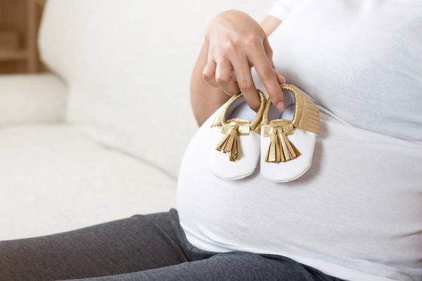 Happy pregnant woman hand holding baby shoes while sitting on gray sofa at home for rest and relax in living room at home. Excited mom when the baby is close to birth and preparing for newborn. - Photo, image