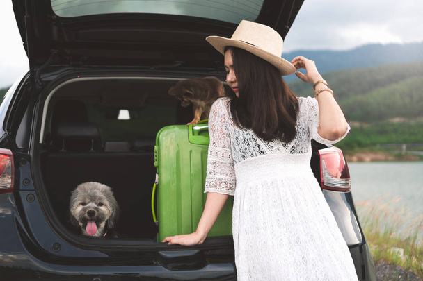 Happy young woman traveler sitting in hatchback car with dogs while parking near lake with mountain view and sunset. Female in white dress enjoys travel with pets in summer vacation in road trips. - Fotó, kép