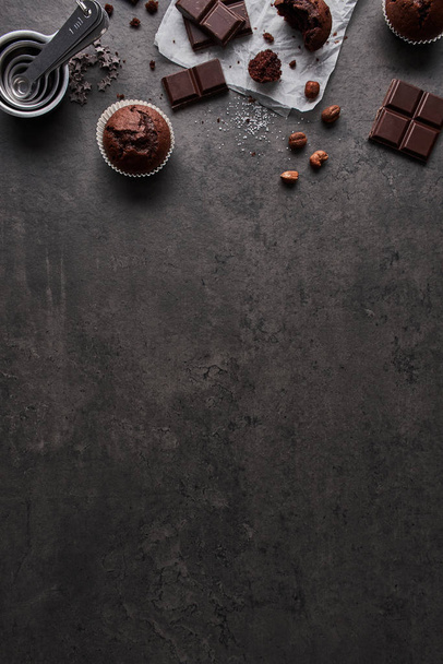 Baking background with chocolate muffins, measuring cups, chocolate, sugar and nuts on dark concrete background. Flat lay with copy space at the bottom. - Фото, зображення