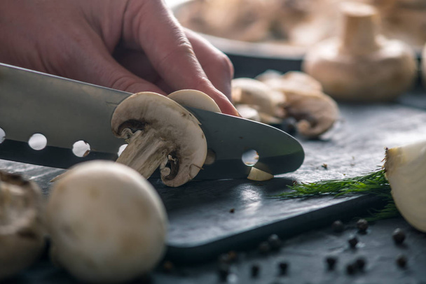 Las manos de las mujeres están cortando champiñones con un cuchillo en la mesa de la cocina oscura. El concepto de cocinar platos de champiñones. De cerca.
 - Foto, imagen