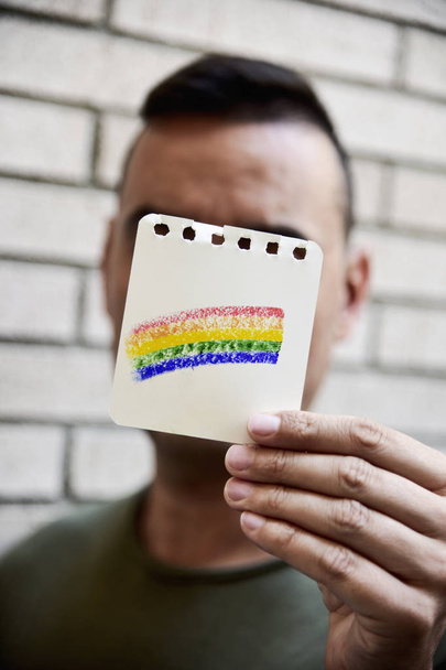 closeup of a young caucasian man holding a piece of paper with a rainbow flag painted in it in front of his face - Photo, Image
