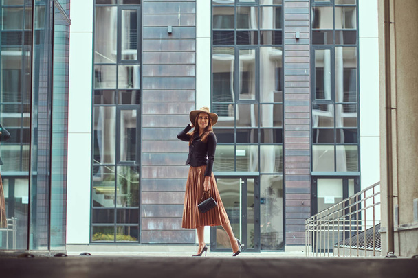 Mujer elegante moda feliz con una chaqueta negra, sombrero marrón y falda con un bolso de embrague caminando por el centro de la ciudad europea
. - Foto, imagen