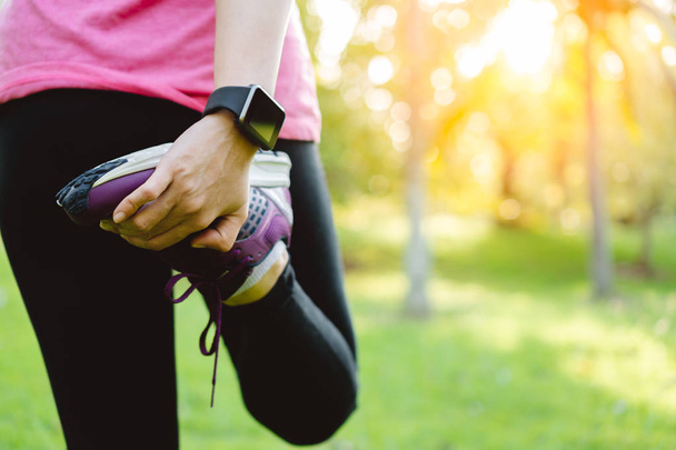 Young runner woman stretching leg before run in park. Close up athletic girl wear running shoes, smart watch warm up outdoor. Sport activity tracker watch at wrist to monitor heart rate during cardio - Foto, Imagen