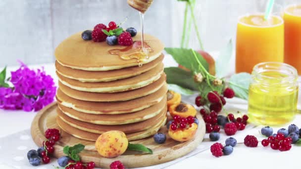 Honey is poured on a stack of delicious pancakes pancakes with summer berries, apricot, peach and mint. Pink phlox bouquet, two glasses of orange juice, honey, light background. - Footage, Video